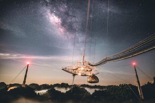 Cables hold a platform in the center of the image, it is night and the sky is filled with stars. The bulge of the Milky Way stretches across the sky in the background.