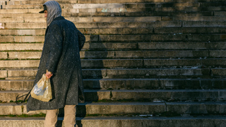 A man in a long coat and hood carries a very expensive shopping bag up some stairs.