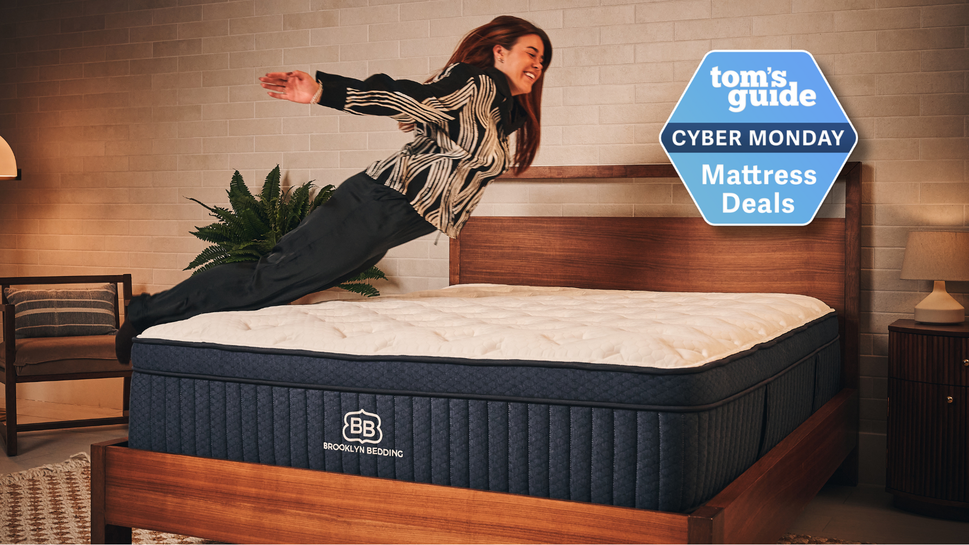 Our mattress tester wearing black and white pyjamas jumping on the Brooklyn Bedding Aurora Luxe mattress on wooden bedframe in warmly-lit sleep studio with Tom&#039;s Guide Cyber Monday mattress deals badge