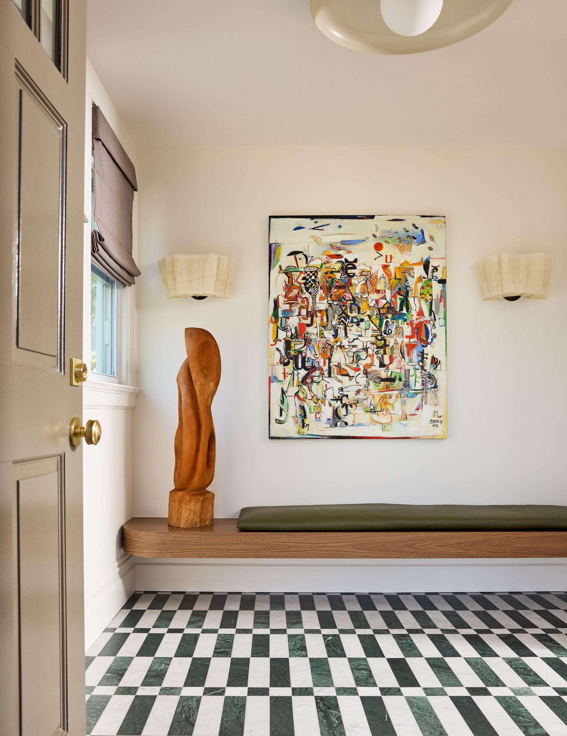 A spacious hallway with a built-in bench seat, sculpture, wall light and artwork above and patterned striped floor tiles. 