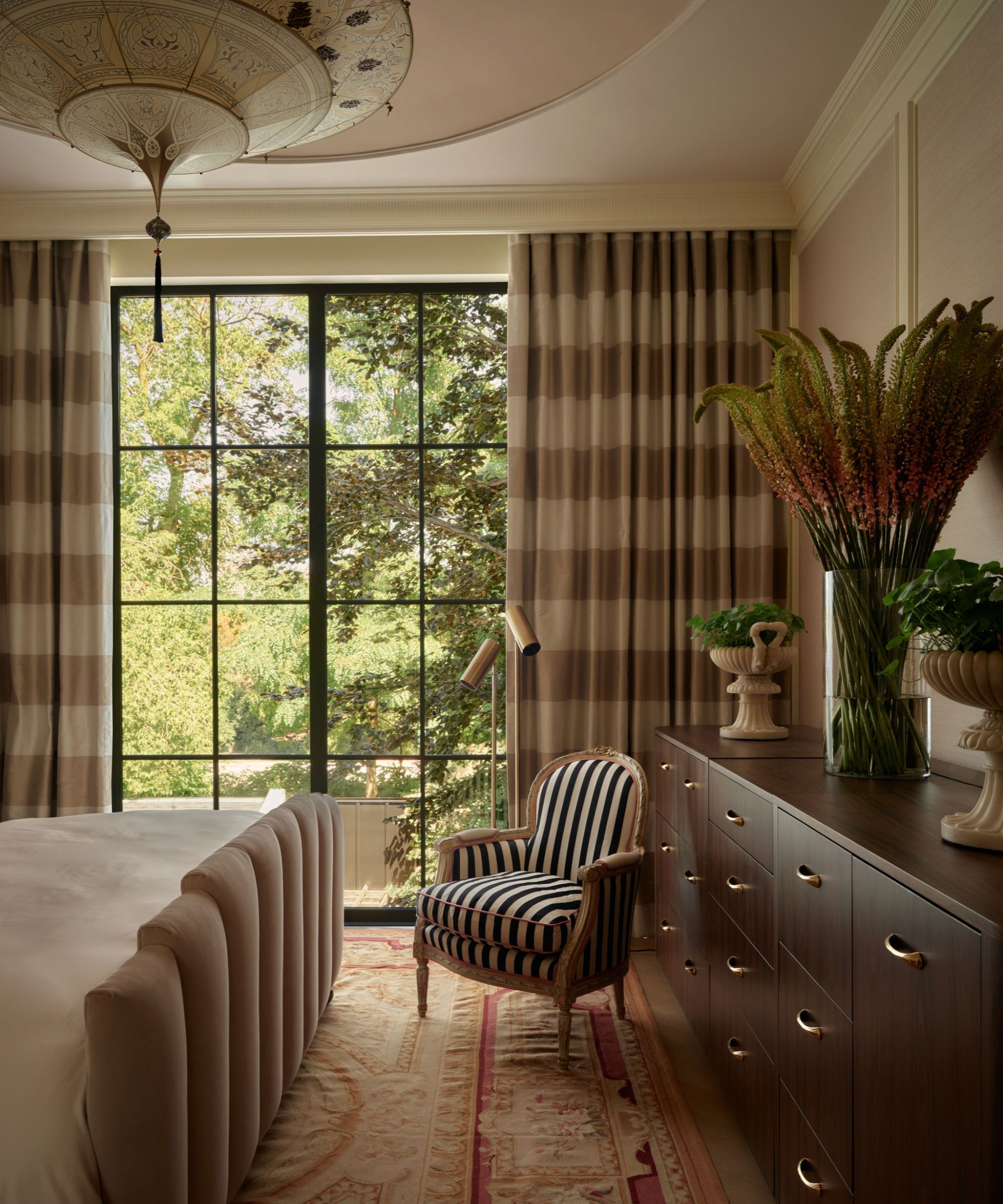 a large soft pink primary bedroom with molding details, large crittal window with beige drapes, a sculptural beige bed, vintage rug, large dark wooden credenza and small striped antique chair