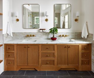 A white bathroom with a rich mid-toned wood bathroom vanity with marble countertops. The backsplash has been created using gray subway tiles and white grout. The brass wall lights with a white shade are from visual comfort