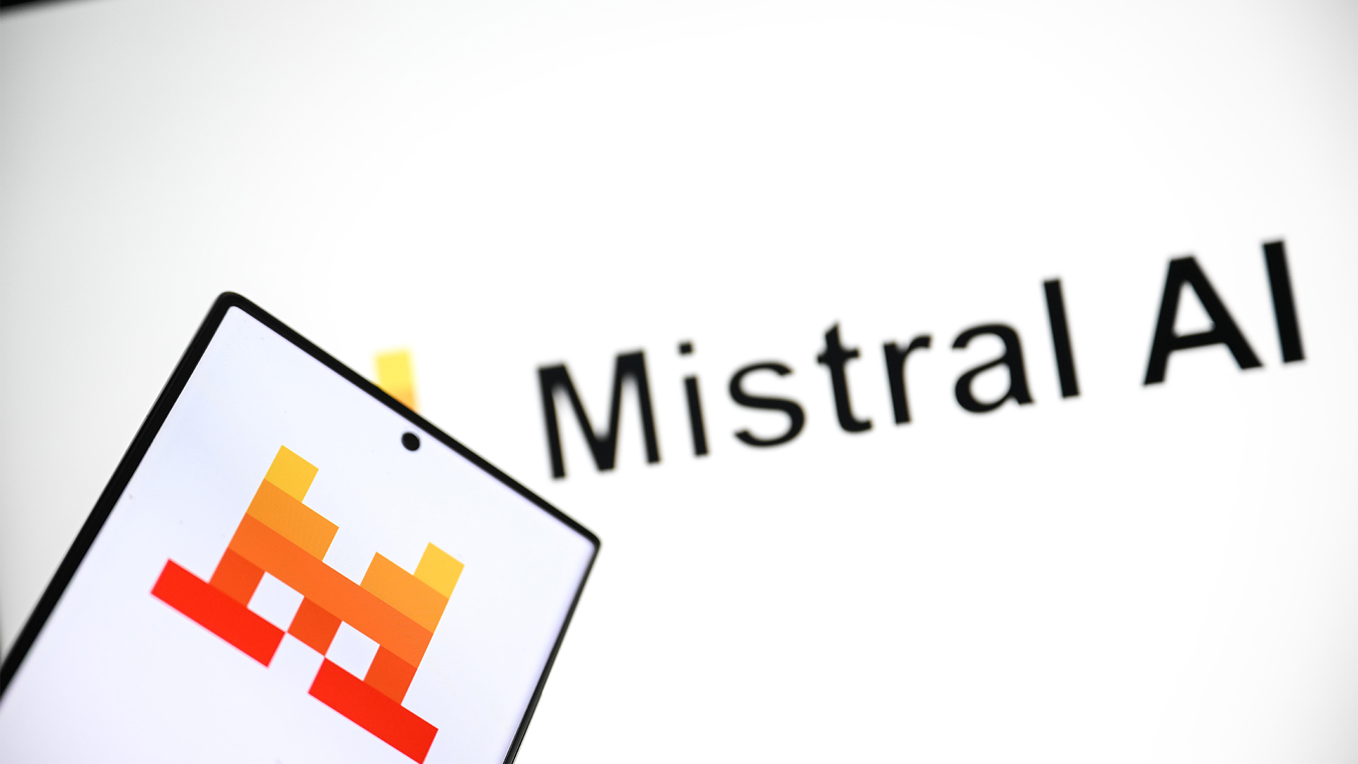 Mistral AI logo and branding pictured on a computer screen, with company logo pictured on smartphone in foreground.
