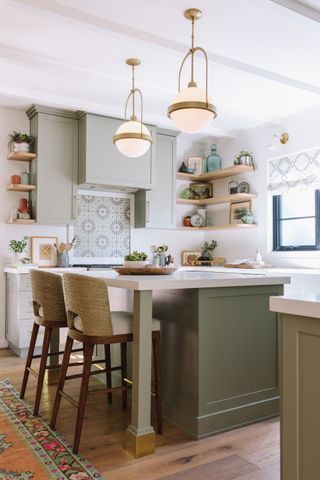 A bright modern kitchen with an island and wicker chairs, open shelving, and sage cabinetry
