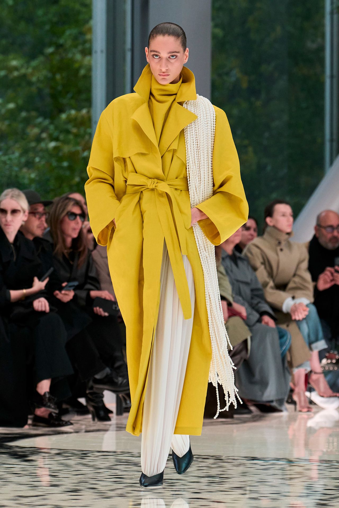 An Ala&iuml;a model wearing the citrus yellow color trend in a yellow trench coat layered over a high-neck yellow blouse styled with white ballon pants, black pointed pumps, and a white fringe scarf.