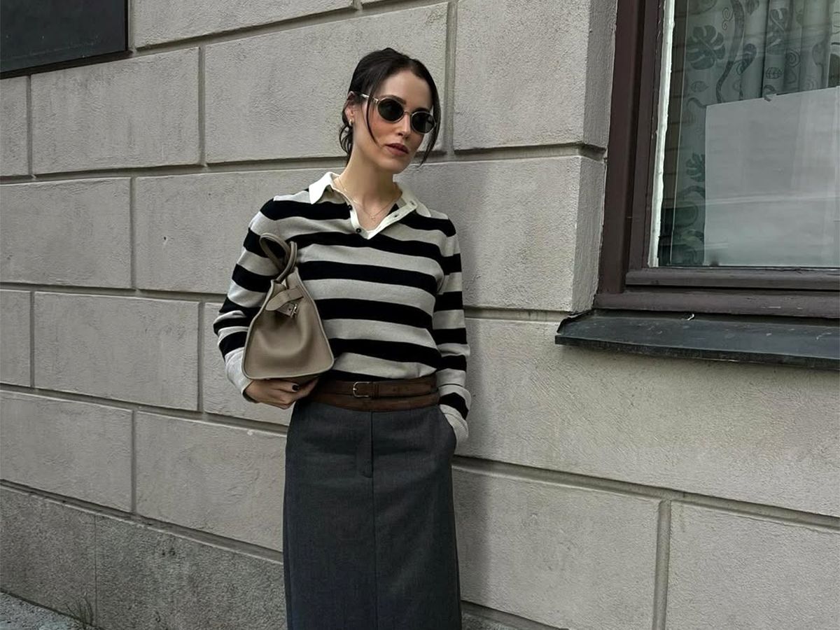 Influencer Anna wears a striped polo sweater and a gray skirt with a brown belt, black loafers, and a light-colored leather bag.