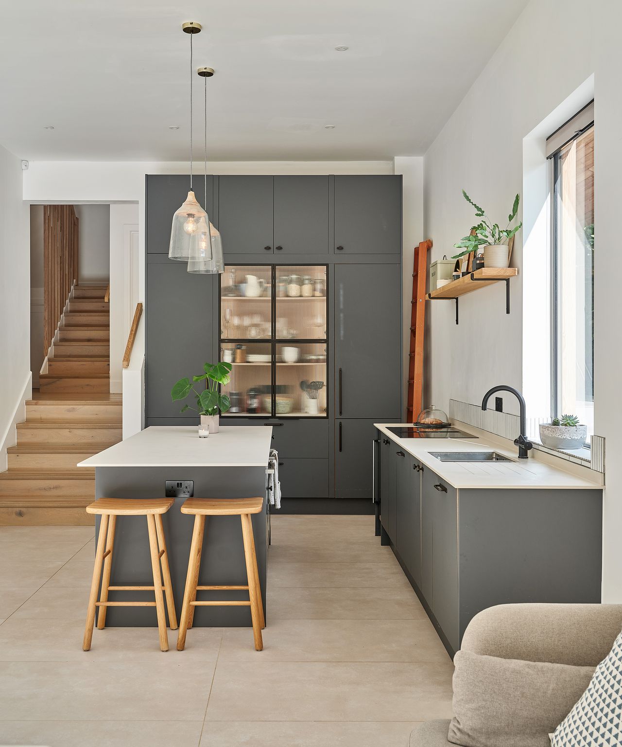 A contemporary kitchen with grey cabinets and white worktops with wooden staircase in the background leading up to another room