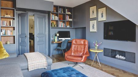 Blue living room with wall-mounted TV, built-in shelving across the back wall and work corner with desktop Apple computer monitor, and tan leather mid-century style armchair