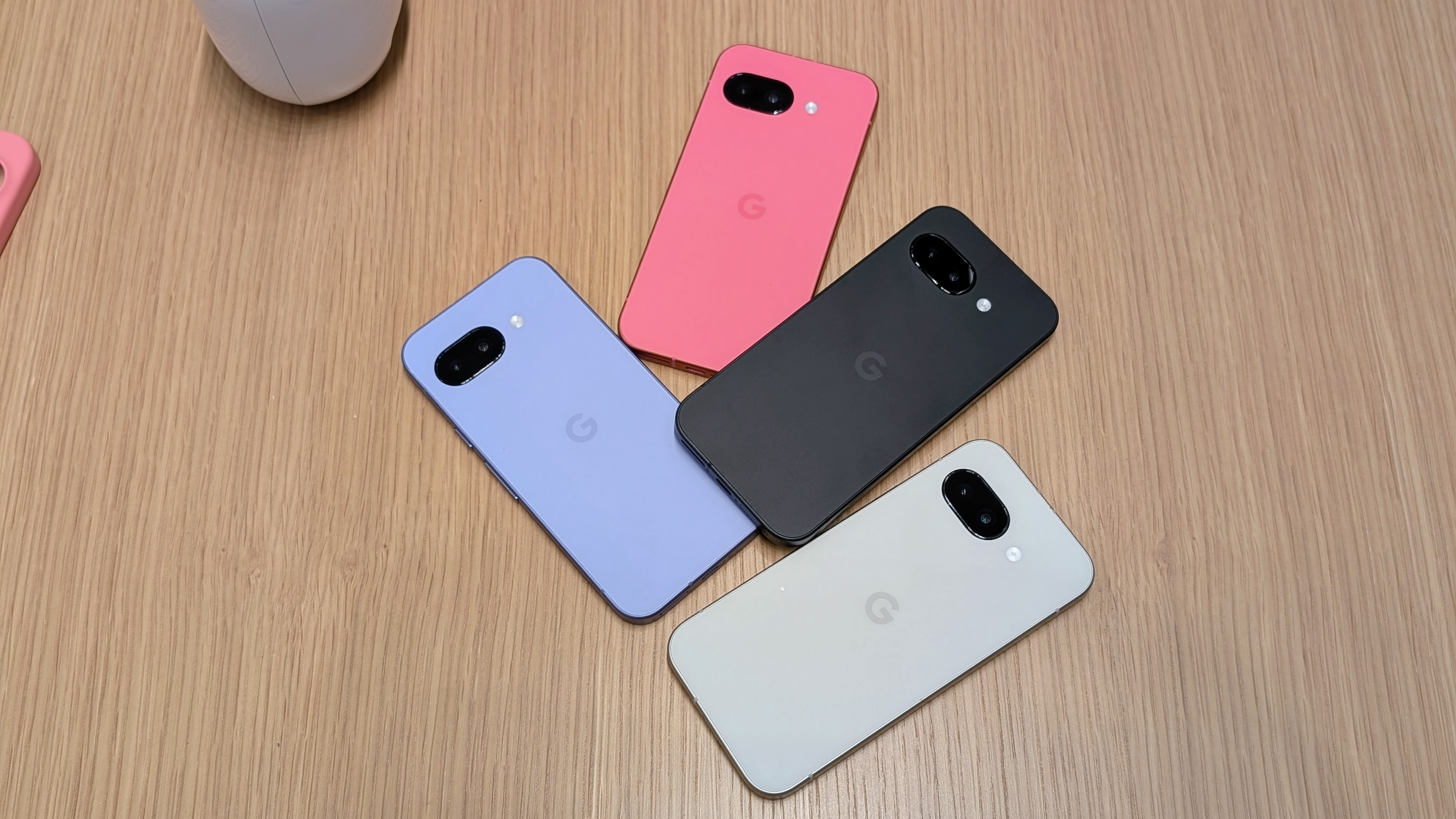 The Iris, Peony, Obsidian, and Porcelain Google Pixel 9a in a jumbled pile on a brown desk surface.