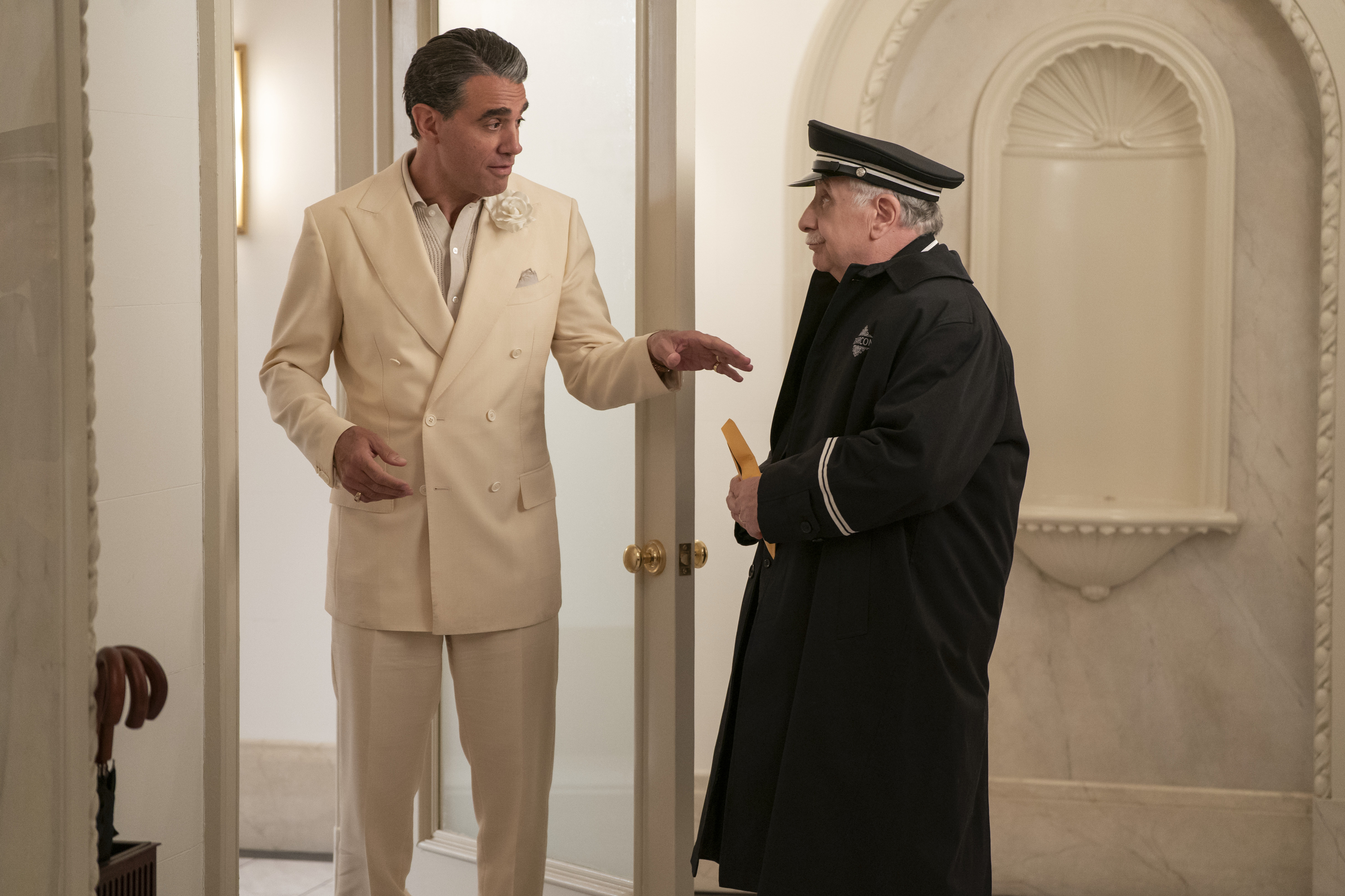 A man (Bobby Cannavale as Nicky Caccimelio) wearing a white suit talks to an older doorman (Teddy Coluca as Lester) holding an envelope as they stand in a building lobby, in &#039;Only Murders in the Building&#039; season 5.