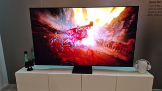 The Samsung S90H OLED TV sits on a credenza in a staged, showroom setting. On the screen is colorful, kinetic, red-tinted imagery.