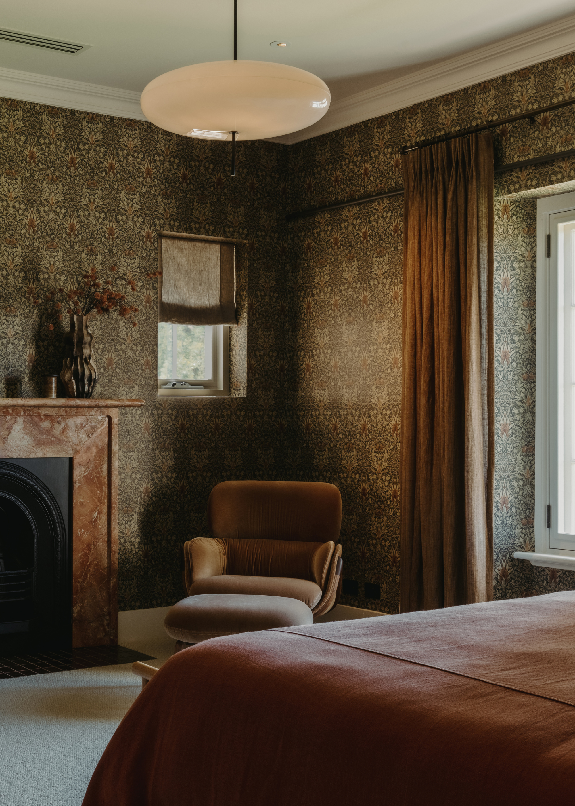 A bedroom with patterned wallpaper, a tan bedscape, a chair with a foot rest, a stone fireplace, a doughnut lamp