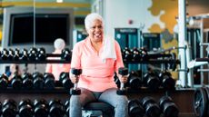 Woman sitting on weight bench in front of rack of dumbbells in gym. She is resting dumbbells on her knees. 