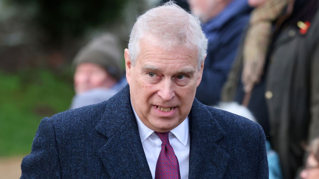 Prince Andrew has white gray hair and wears a large overcoat with a white shirt and red tie underneath