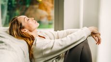 An exhausted woman sits on the floor and leans against a bed. Her eyes are closed. 