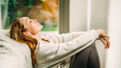 An exhausted woman sits on the floor and leans against a bed. Her eyes are closed.