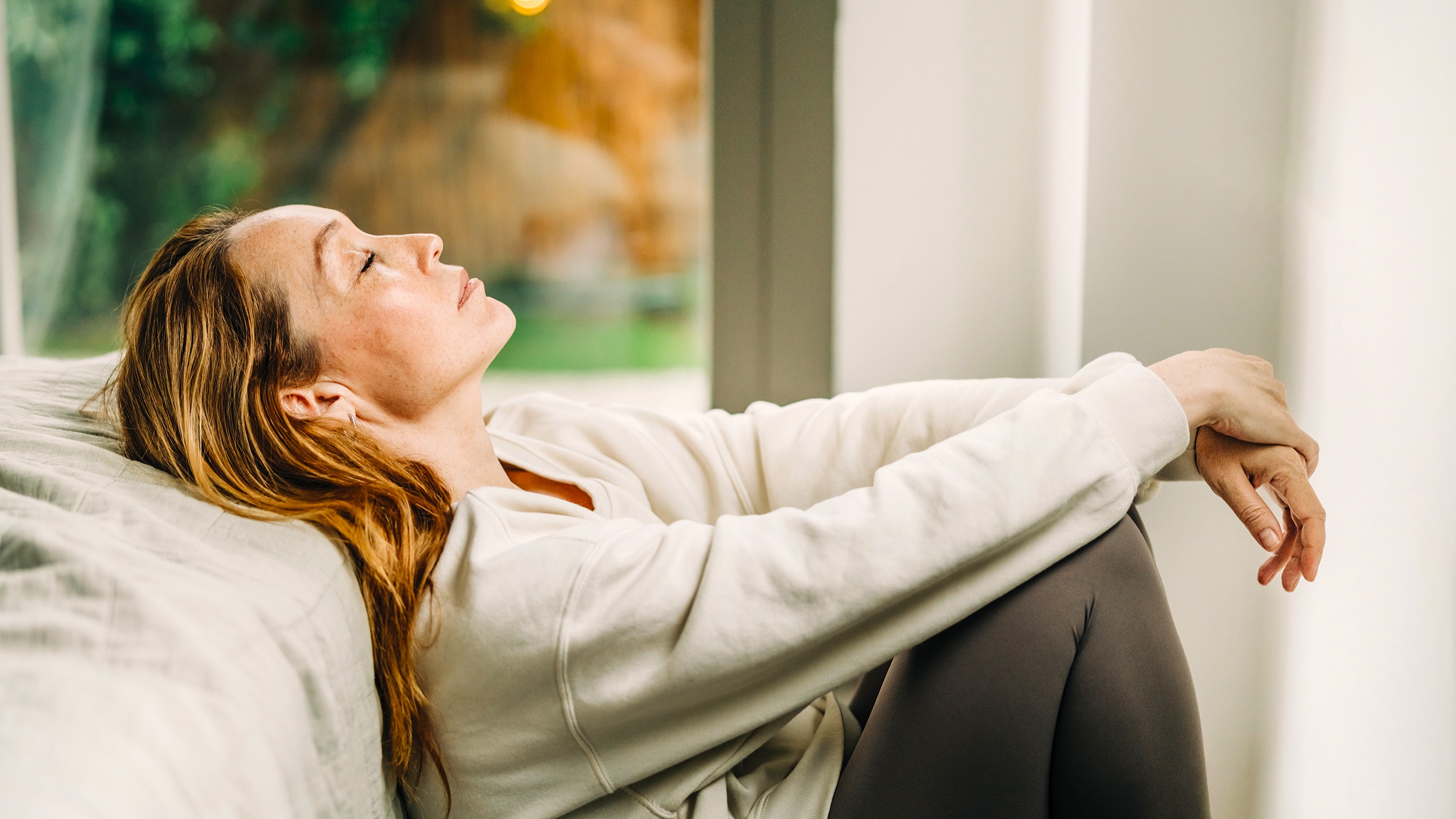 An exhausted woman sits on the floor and leans against a bed. Her eyes are closed. 