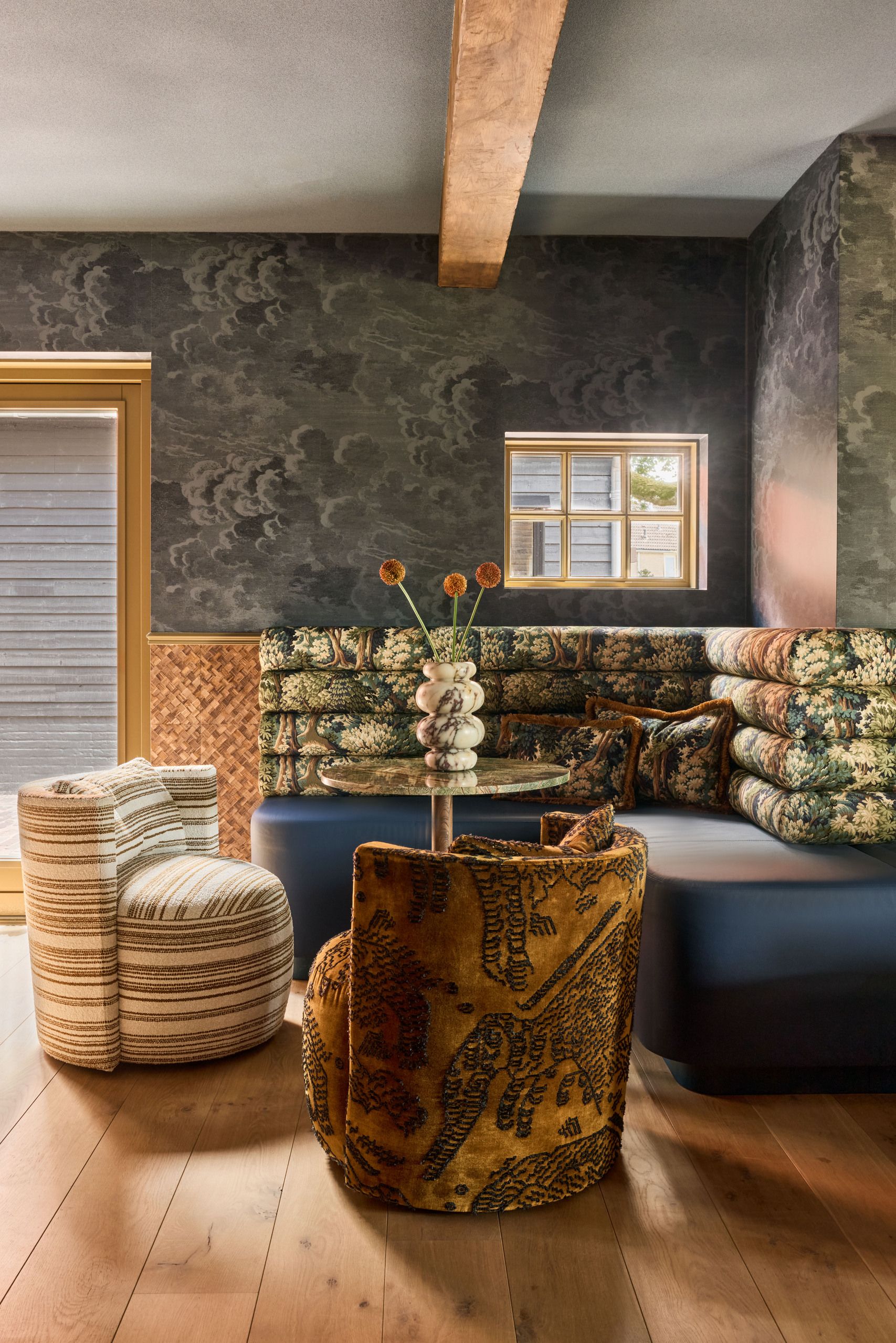 A corner dining bench area with a blue seat and patterned back. There are two patterned chairs opposite the bench and blue wallpaper on the walls.