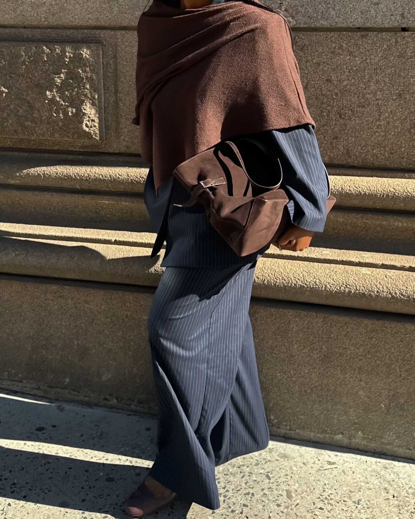 A woman wears a brown scarf, blue blazer, blue pants, and a brown suede tote.