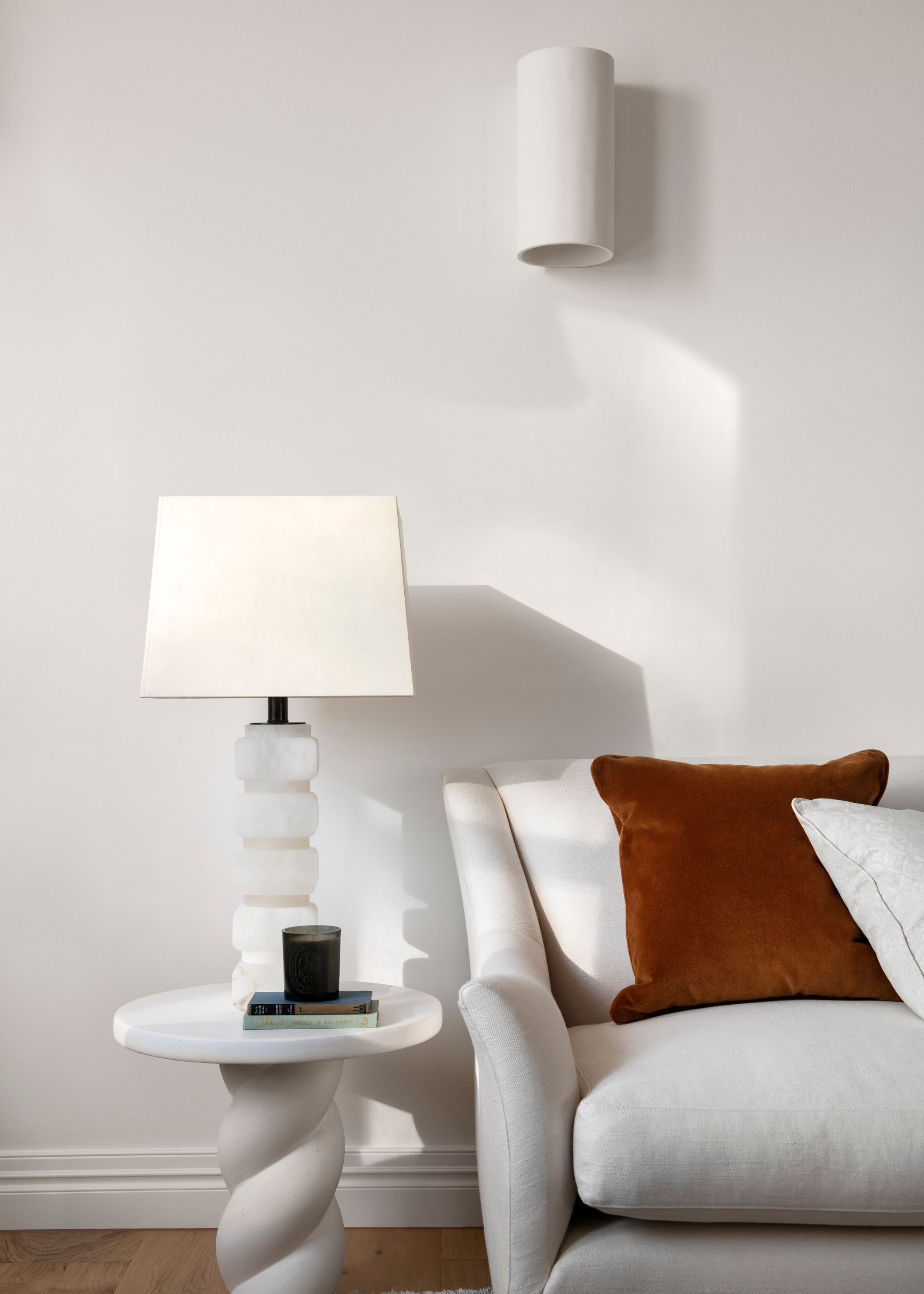A white living room with a sofa and a side table with a lamp on top. On the table are also some books with a candle on top. On the couch is a deep red cushion to add contrast to the space.
