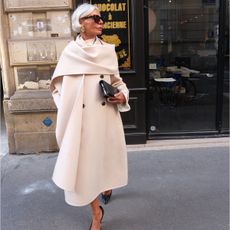 Grece wears a long ivory coat with an attached scarf and a black leather clutch.