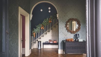 Festive wallpapered entryway with wooden parquet flooring, a circular mirror, a black chest of drawers, and a white staircase in the background with a festive garland and ribbons.