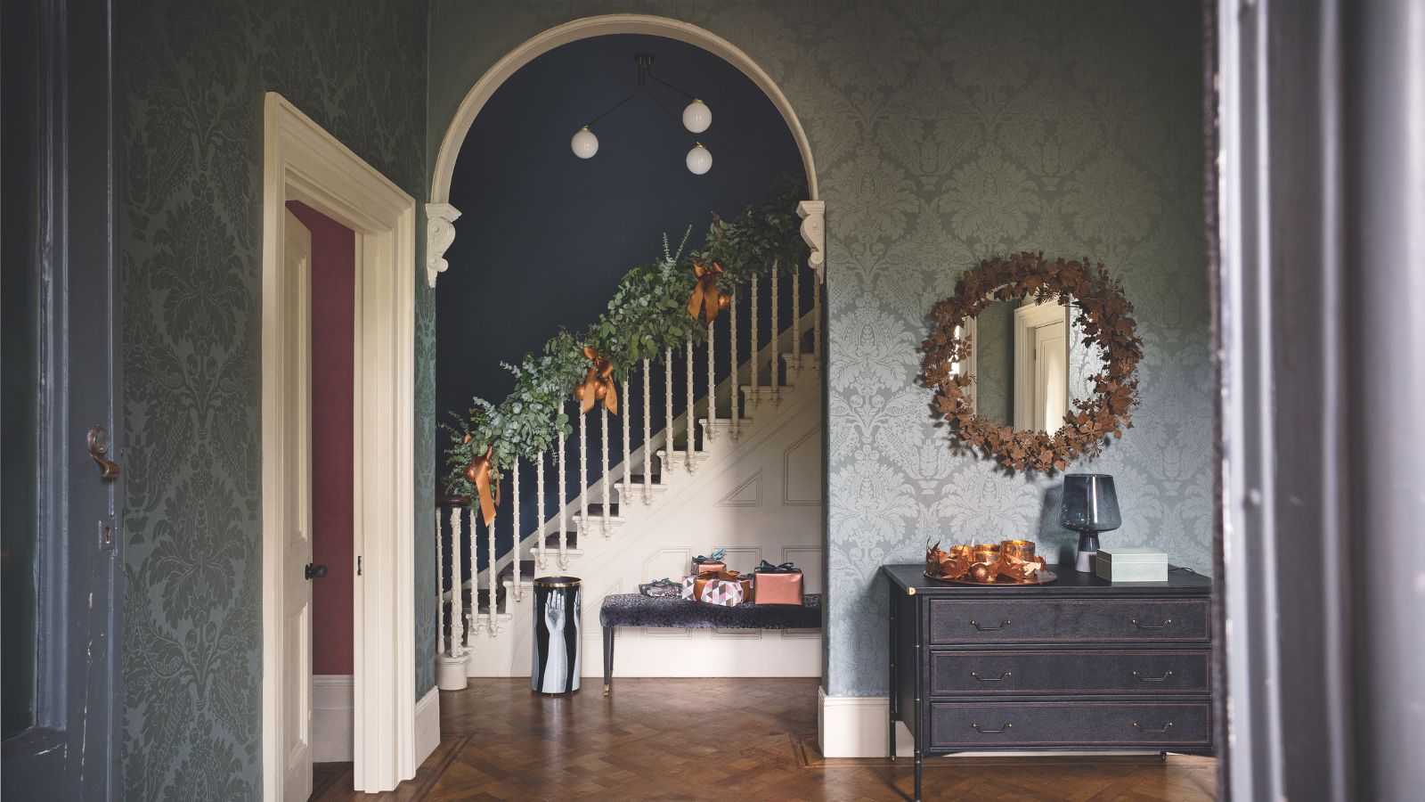 Festive wallpapered entryway with wooden parquet flooring, a circular mirror, a black chest of drawers, and a white staircase in the background with a festive garland and ribbons.