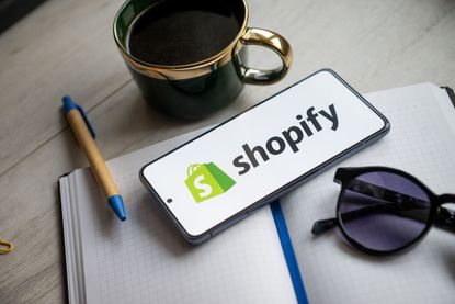 Shopify logo on a smartphone that&#039;s placed on a table next to a pen, notebook and cup of coffee