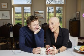 Sarah Beeny and her husband Graham Swift sit at their dining table, each holding a mug that is resting on the table. They are both looking into each other's eyes and smiling.
