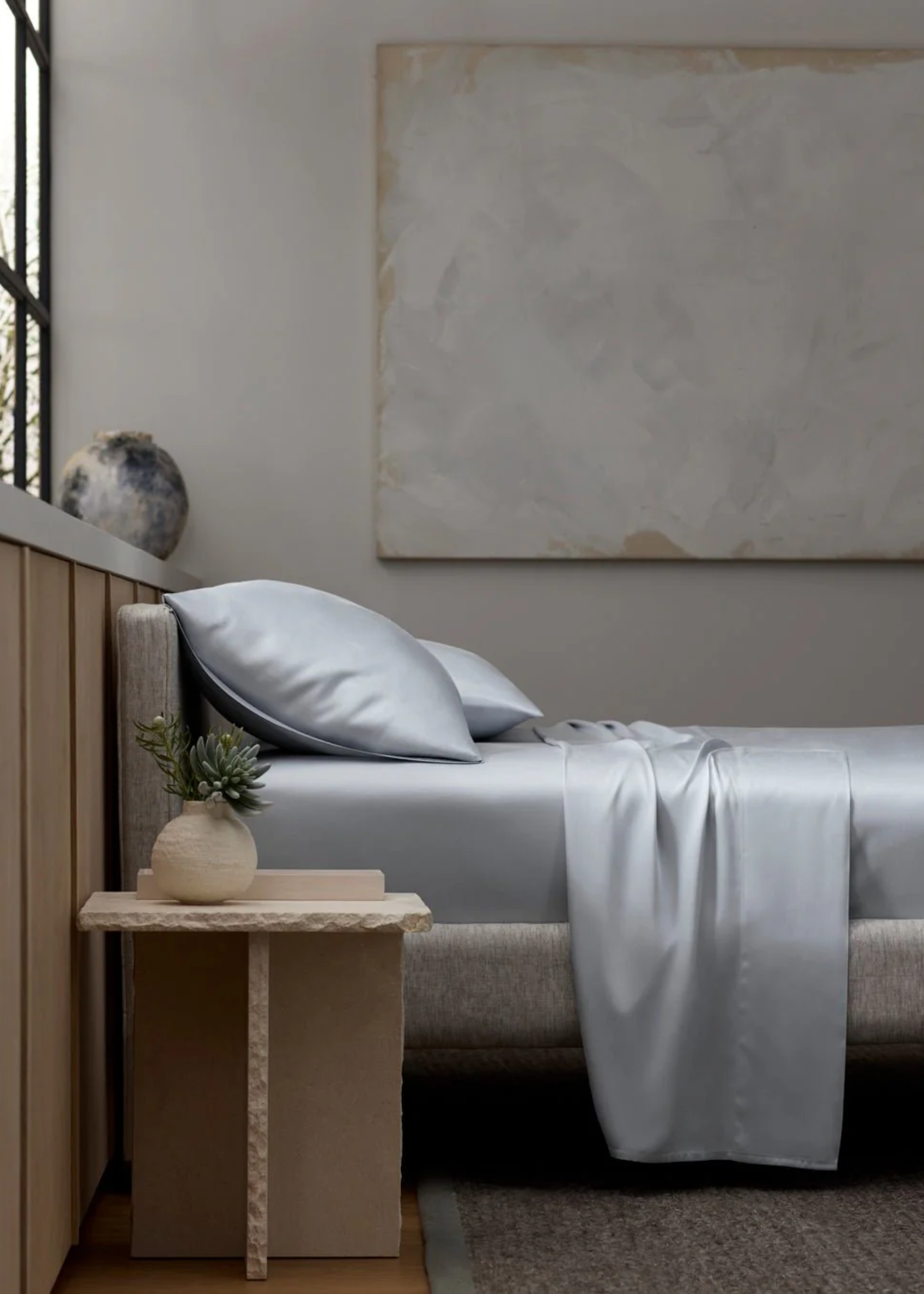 A minimalist bedroom with a structured side table a vase with green stems, a large rug, an unframed painting, and a bed with pale blue bedding