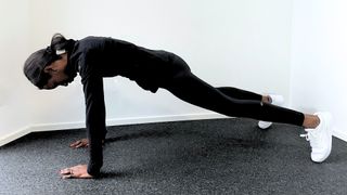 Fitness trainer Dilan Gomih performs a plank in an empty room. She is close to the ground, with her arms straight and hands flat on the floor. The only other point of contact with the floor is her toes. Her body forms a straight line and her gaze is directed at the floor. She is wearing a long-sleeved sports top, leggings and sneakers.