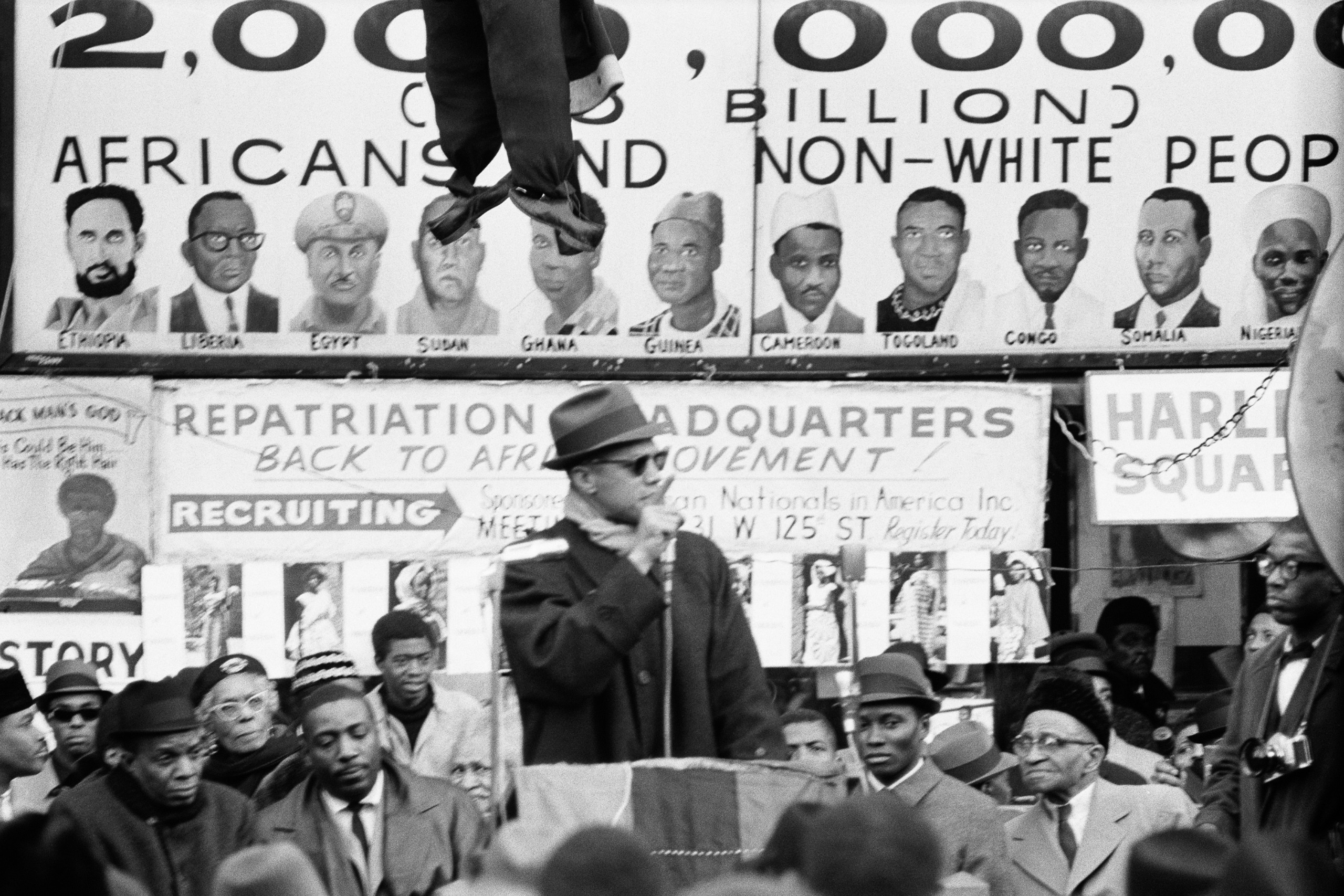 Malcolm X speaks at a podium in Harlem, one finger raised, in front of banners advertising a "Back to Africa" repatriation movement meeting