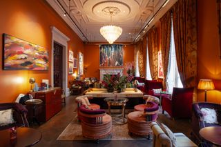 A color-drenched room with orange walls is filled with lavish furniture, including Art Deco-style banquettes and armchairs in velvet, Murano glass chandeliers, abstract paintings, and more.