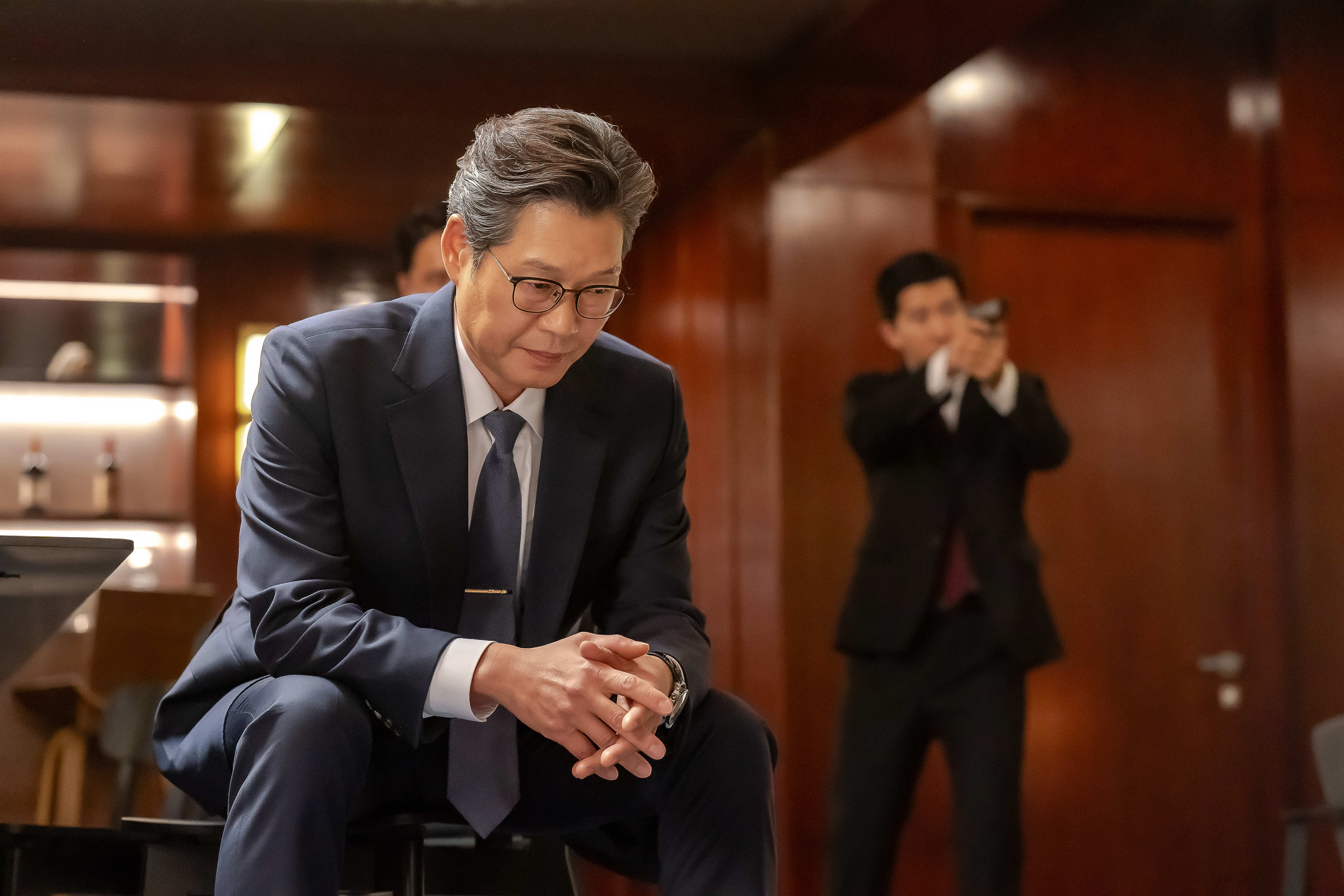 Yoo Jae-myung wears a sit down and looks contemplative while another man holds up a gun behind him in &#039;Tempest.&#039;