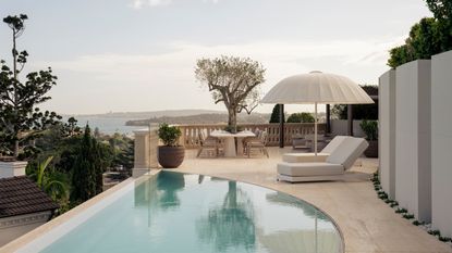 Image of a crescent-shaped pool on a deck that overlooks an ocean view. There are white lounge chairs with a white umbrella. There is a glass partition by the balcony edge.