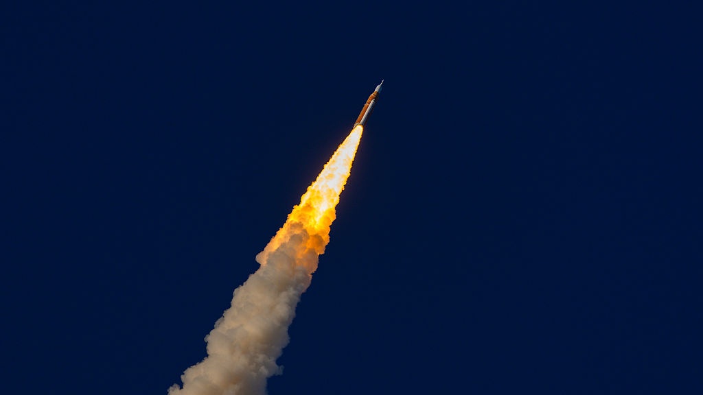 A rocket that's orange against the blue background of the sky is launching.