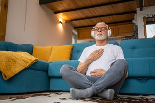 Mature senior man sitting on floor practicing guided meditation at home, relaxing body and mind concept