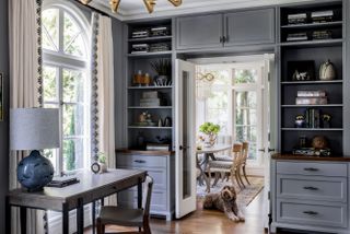 writing desk with blue lamp by window with built in grey storage cabinets around and above a double door with view to dining space beyond