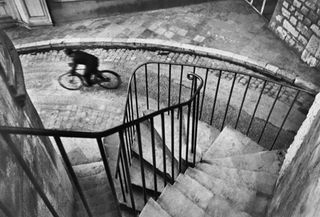 Black and white 1932 French image, stone spiral steps with black metal guide rail, stone road below, action shot of man on a bicycle, stone walls and narrow cobbled street