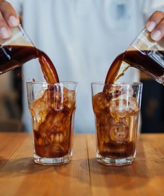 A person wearing a light blue shirt pouring coffee into two glass cups with ice cubes in, stood on a rich wooden surface