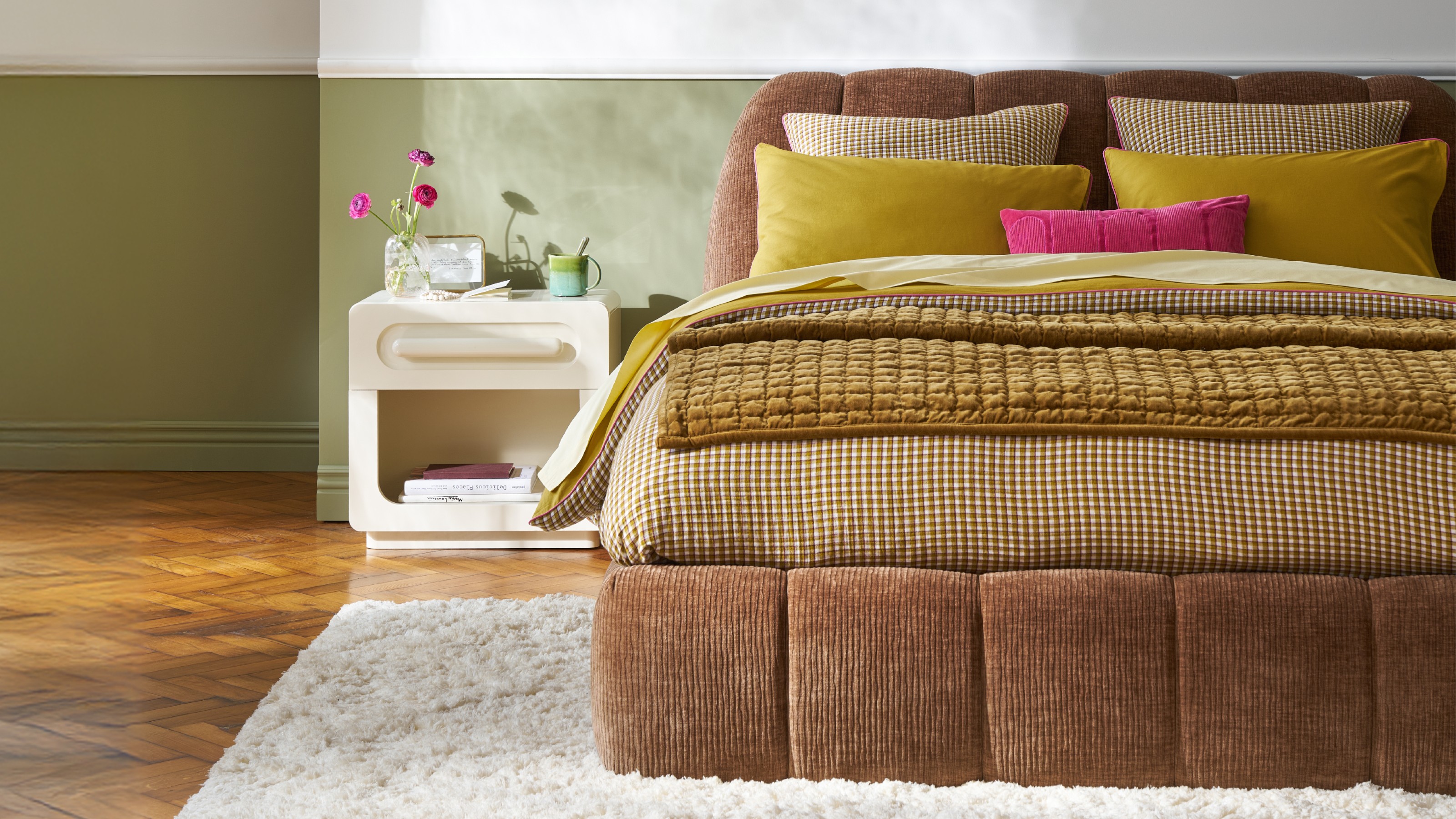 A bedroom with a brown upholstered bed dressed with gingham bedding and a lacquered ivory-coloured bedside table next to it
