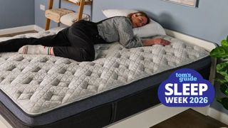 The image shows a light haired woman wearing a black and white gingham shirt, black dungarees and white bed socks lying on her side on a white and blue mattress in a neutrally decorated bedroom. Overlaid on top in the bottom right hand corner a blue cloud-shaped badge that reads Tom's Guide Sleep Week 2026.