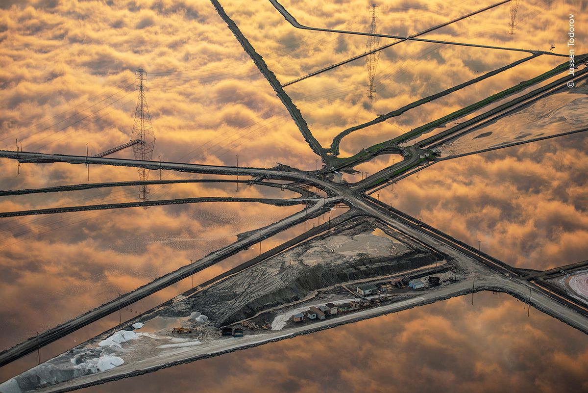 Aerial view of intersecting roads amidst industrial salt ponds reflecting a fiery sky. Power lines traverse the scene, creating a geometric pattern