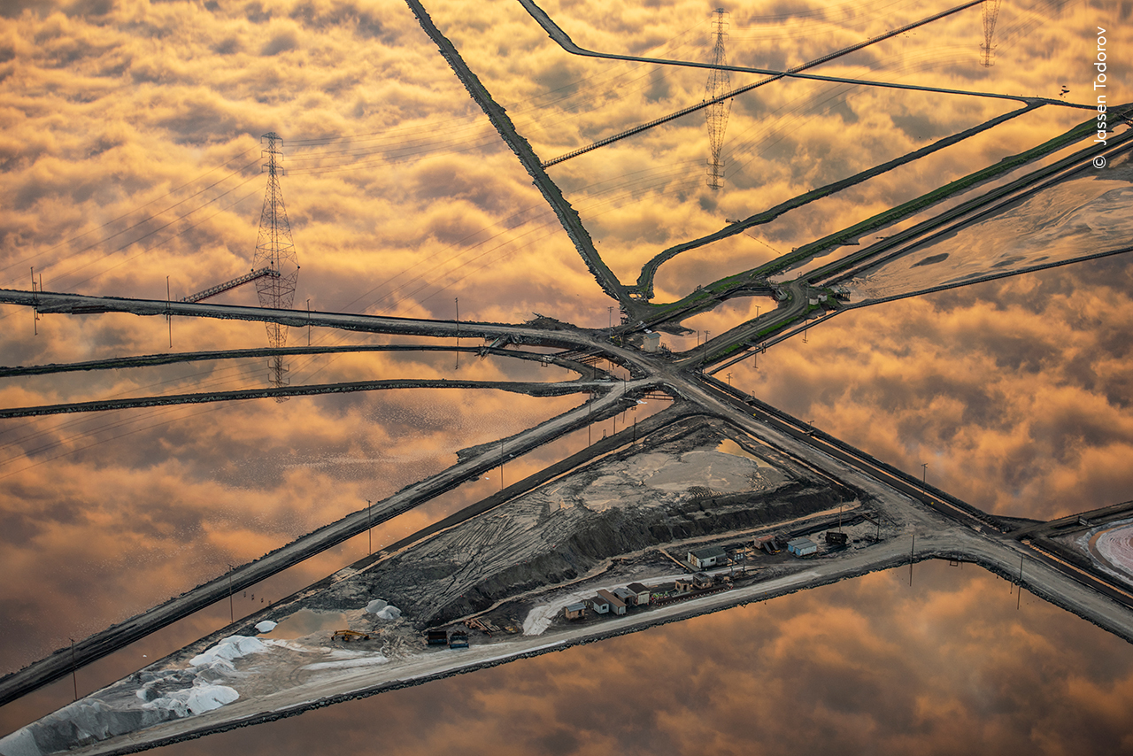 Aerial view of intersecting roads amidst industrial salt ponds reflecting a fiery sky. Power lines traverse the scene, creating a geometric pattern