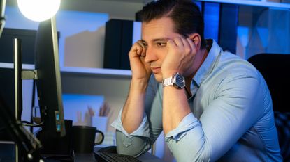 A man sitting at his computer desk on his desktop with his head in his hands, looking a little frustrated.