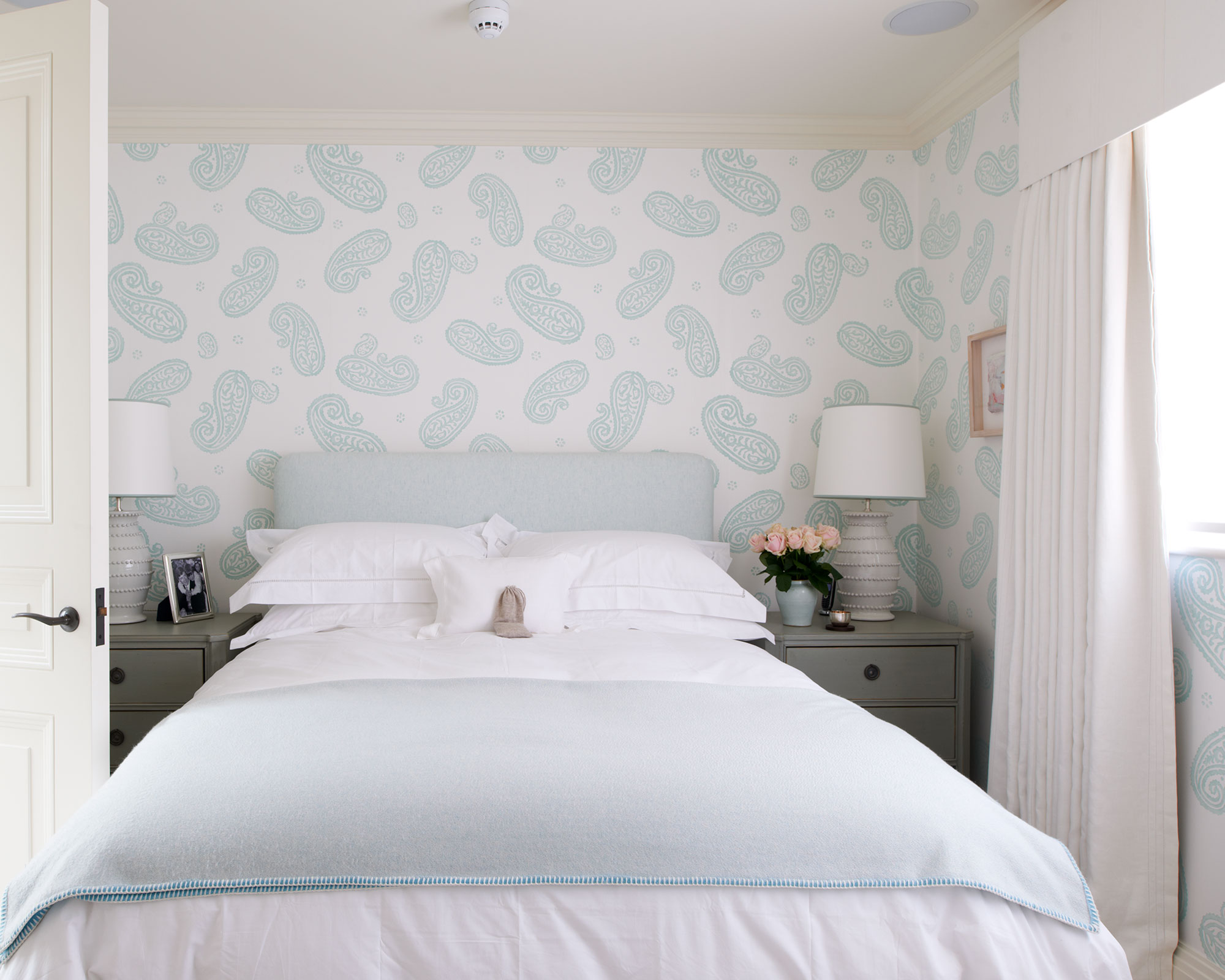 A small Thorp bedroom with pale blue wallpaper and headboard and white drapes.