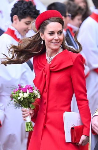 Kate Middleton wearing a red coat and holding flowers tossing her hair in the wind on Commonwealth Day 2025