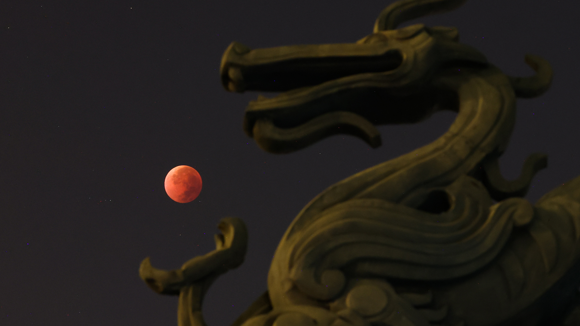 A blood red moon is positioned against the dark sky to appear as if it is held in the claw of a stone dragon statue in the foreground.