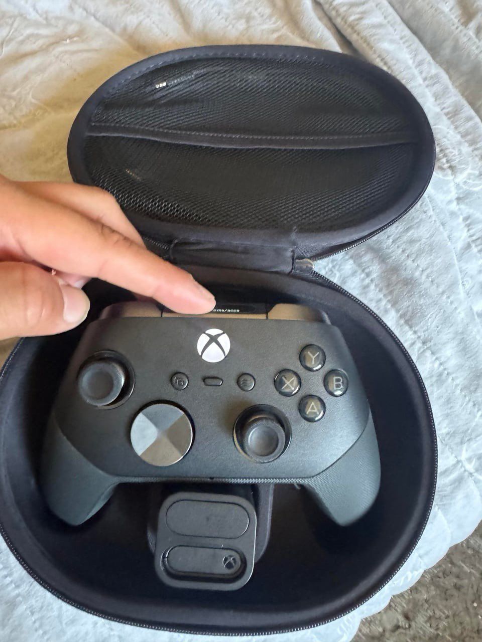 A black Xbox Elite wireless controller sits inside a round, zippered carrying case, featuring a metallic D-pad and standard gaming button layout.