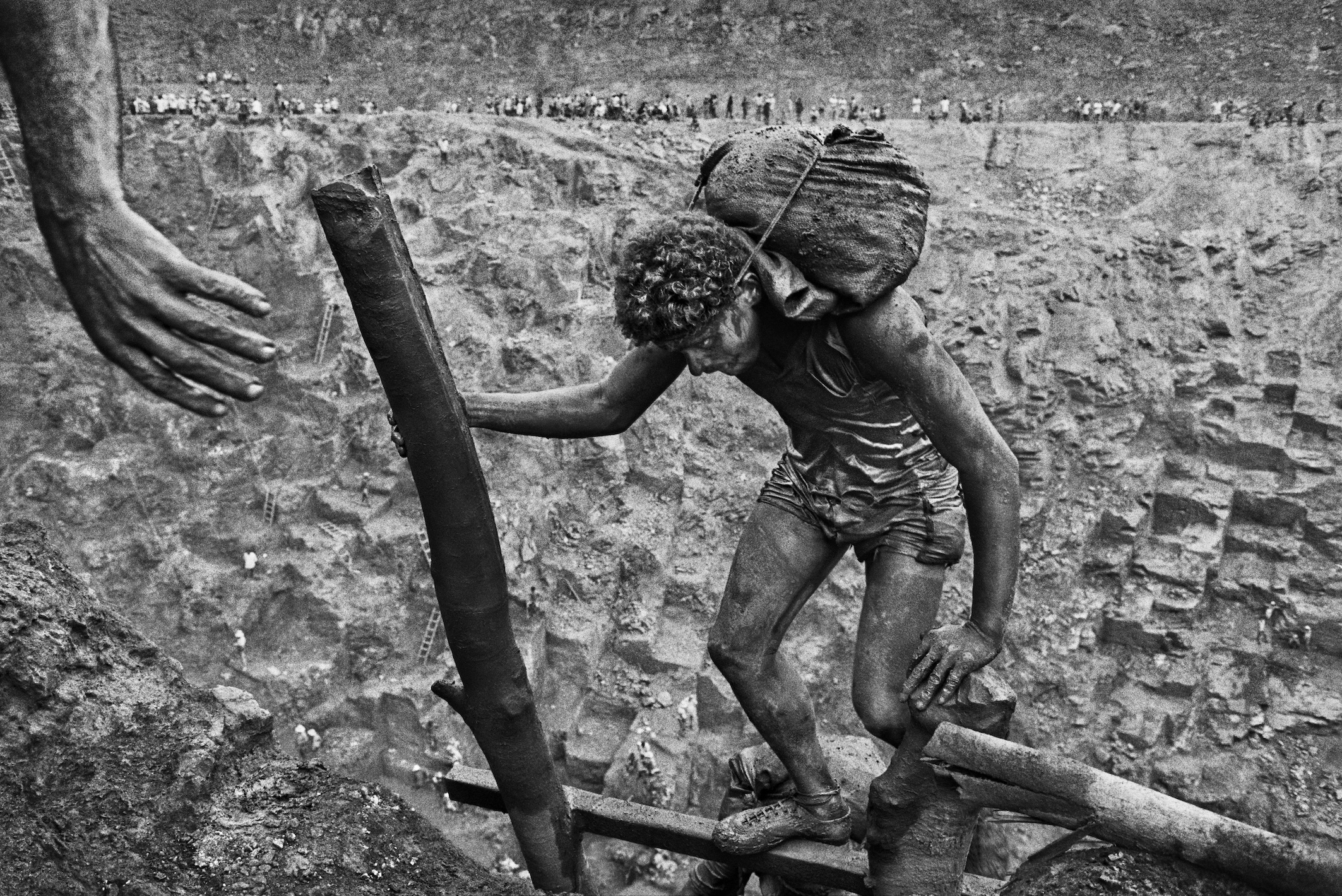 In a stark, black-and-white image of a muddy gold mine, a young male laborer struggles up a wooden plank while carrying a heavy, sack-like load on his back, as a large, helping hand reaches in from the upper left.
